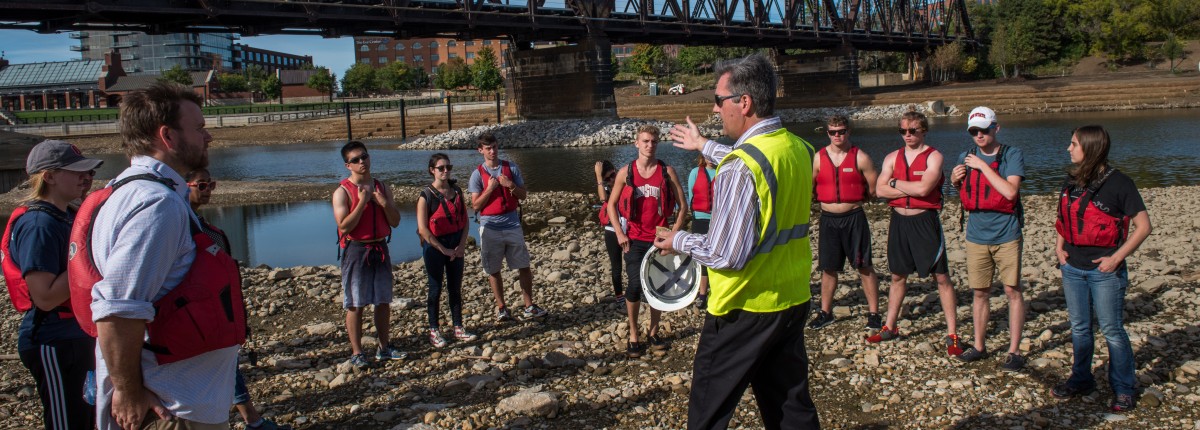 a speaker talks to a group of students beside Olentangy River preparing for canoe trip
