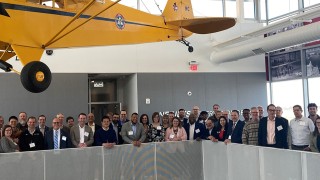2023 Sustainable Aviation Forum attendees pose under a suspended aircraft at the OSU Airport.
