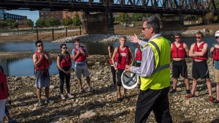a speaker talks to a group of students beside Olentangy River preparing for canoe trip